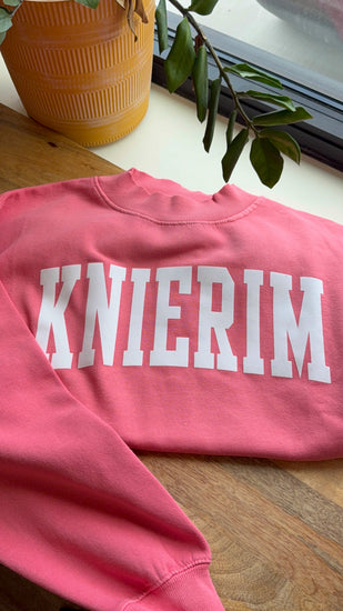 Pink sweatshirt with 'KNIERIM' text on a wooden surface with a plant in the background
