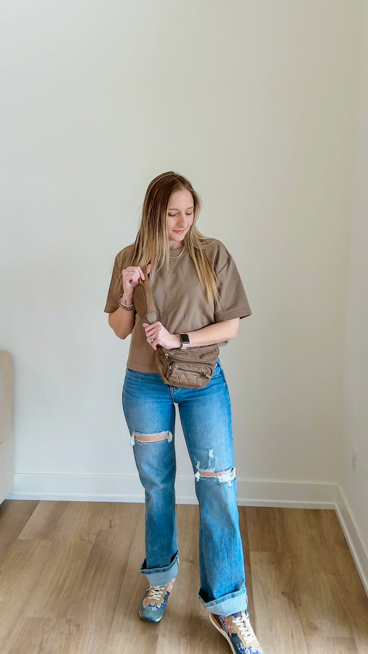 Woman wearing a brown oversized shirt and blue jeans standing in a room with a plain wall.