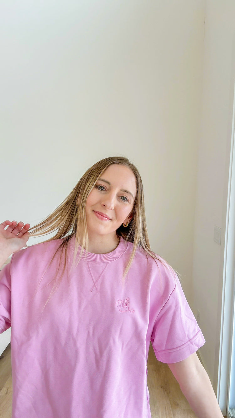 Person wearing a pink t-shirt with a monogram, standing against a plain wall.